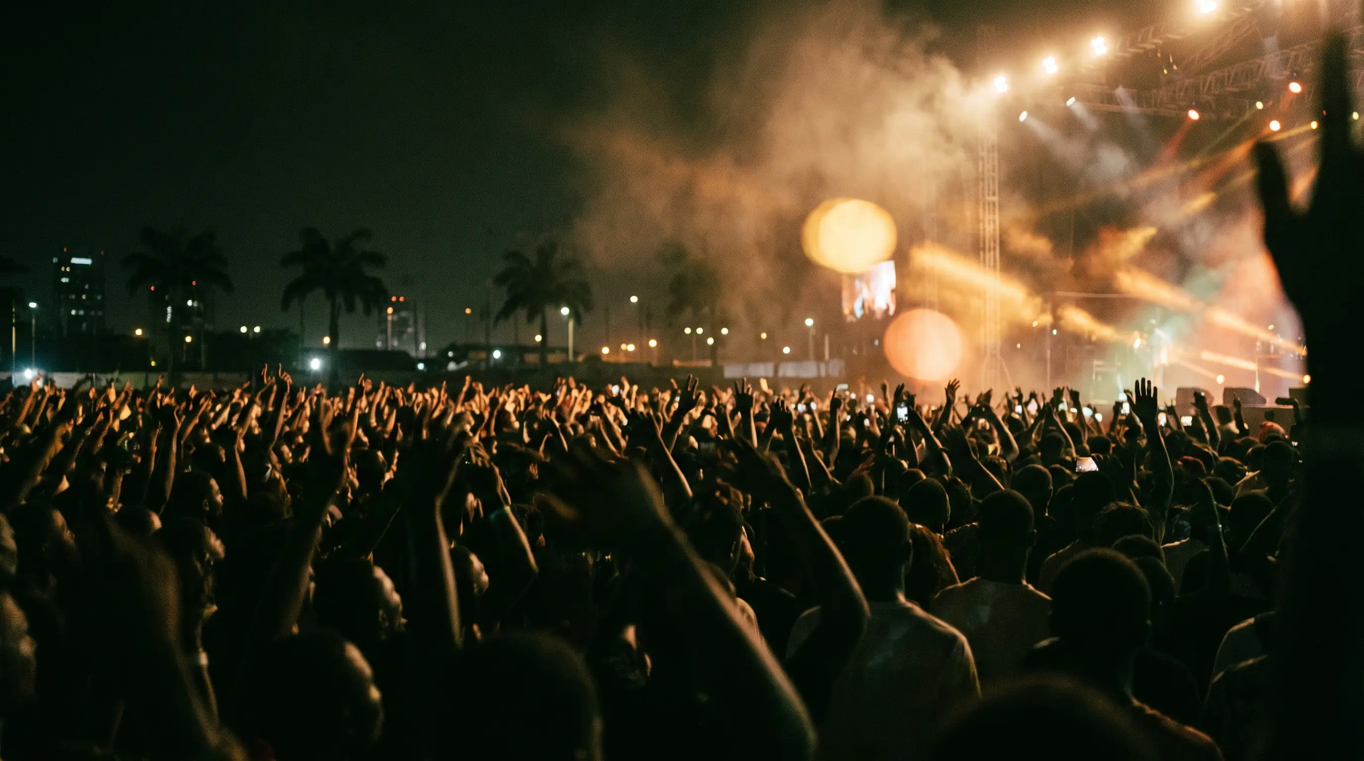 A packed night crowd with hands and phones raised, stage lights and haze above them, palm trees and city lights on the horizon.