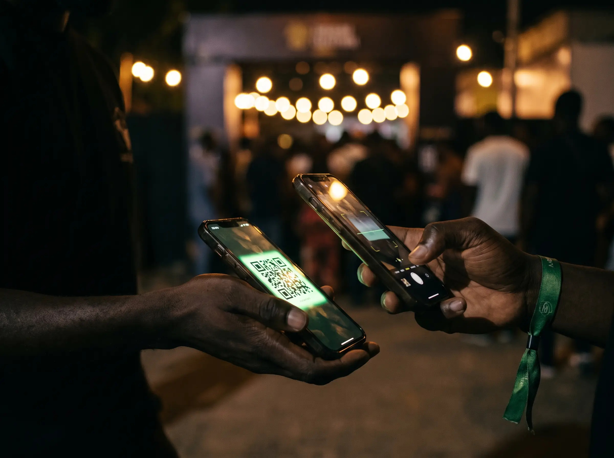 Two hands at a venue entrance at night, one holding a phone showing a ticket QR code, the other holding a phone scanning it, with the lit doorway and queue behind.
